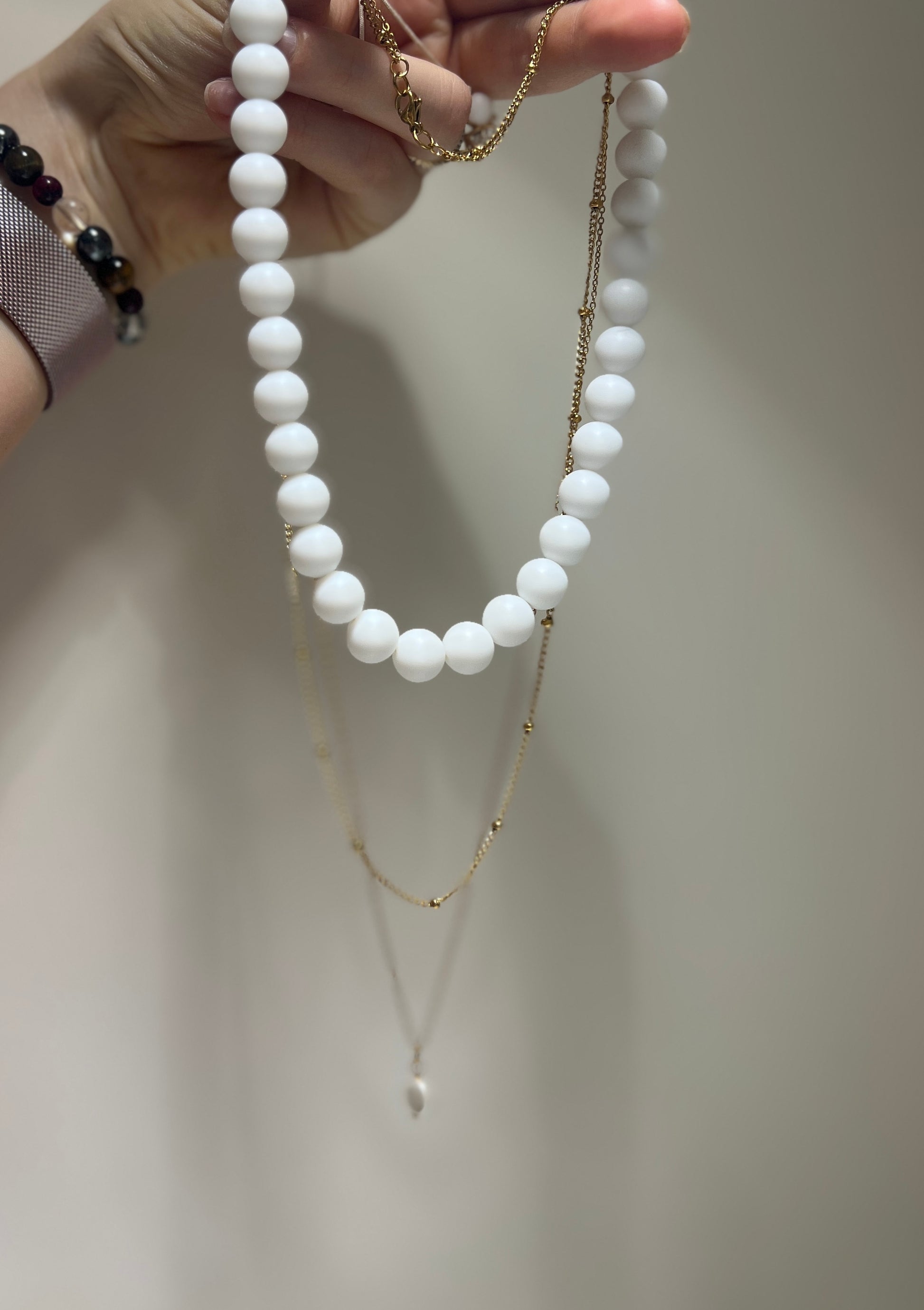 White beaded necklace and gold chain held by a hand against a neutral background