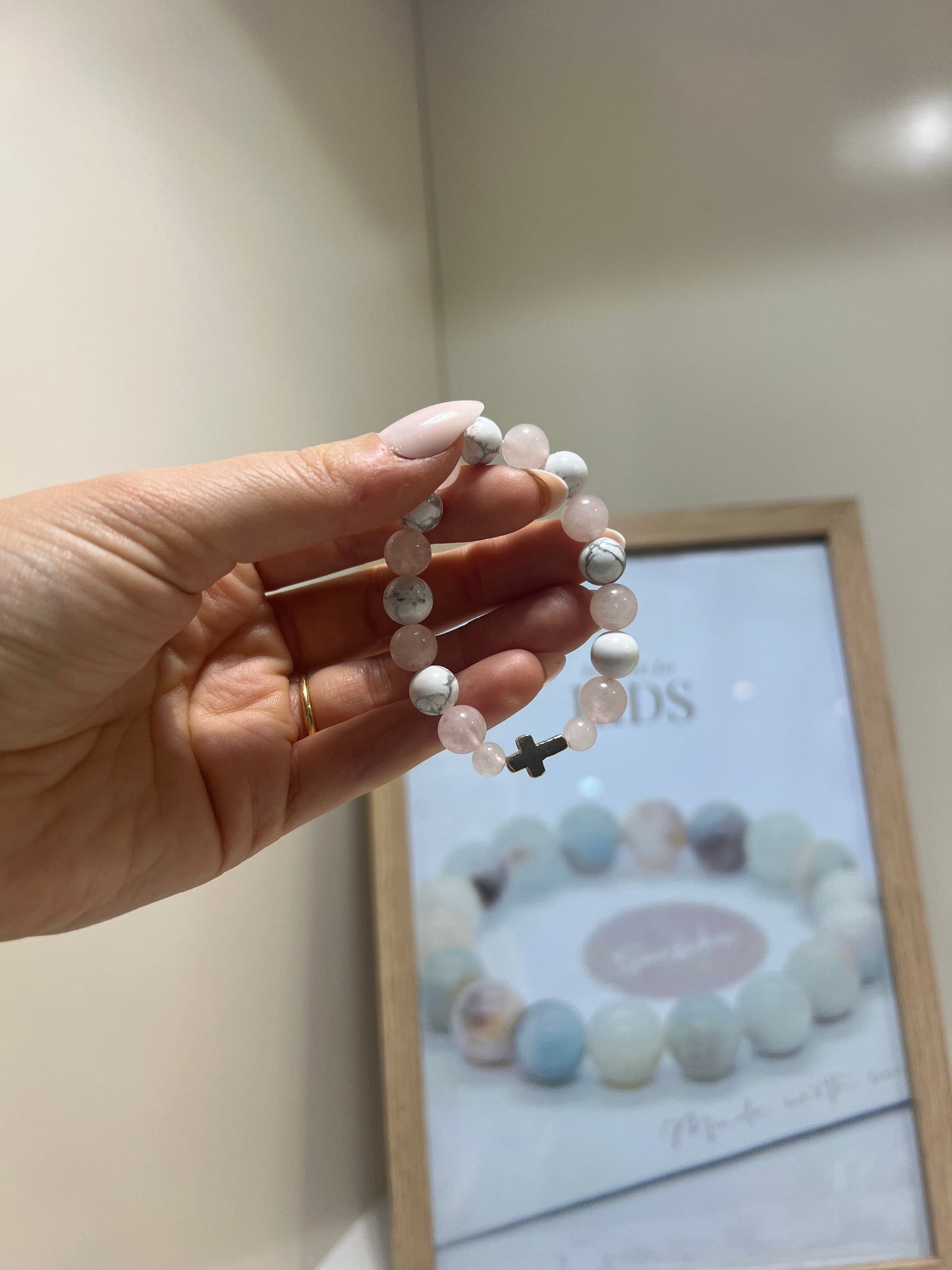Hand holding a beaded bracelet with a cross charm in front of a framed picture.